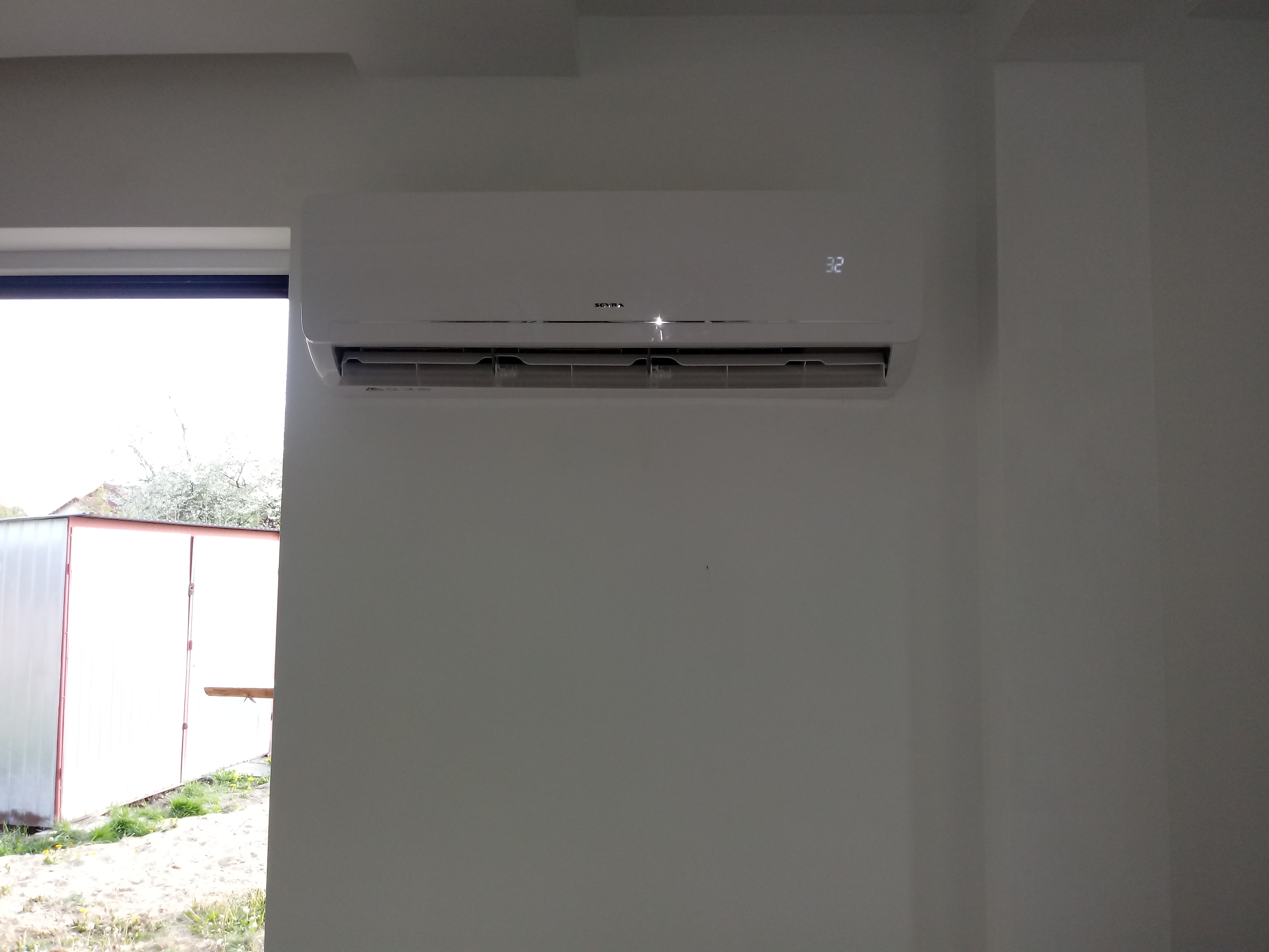Biały klimatyzator marki Seyra zamontowany na białej ścianie, z wyświetlaczem pokazującym temperaturę 32 stopnie Celsjusza, widok na zewnątrz przez okno.