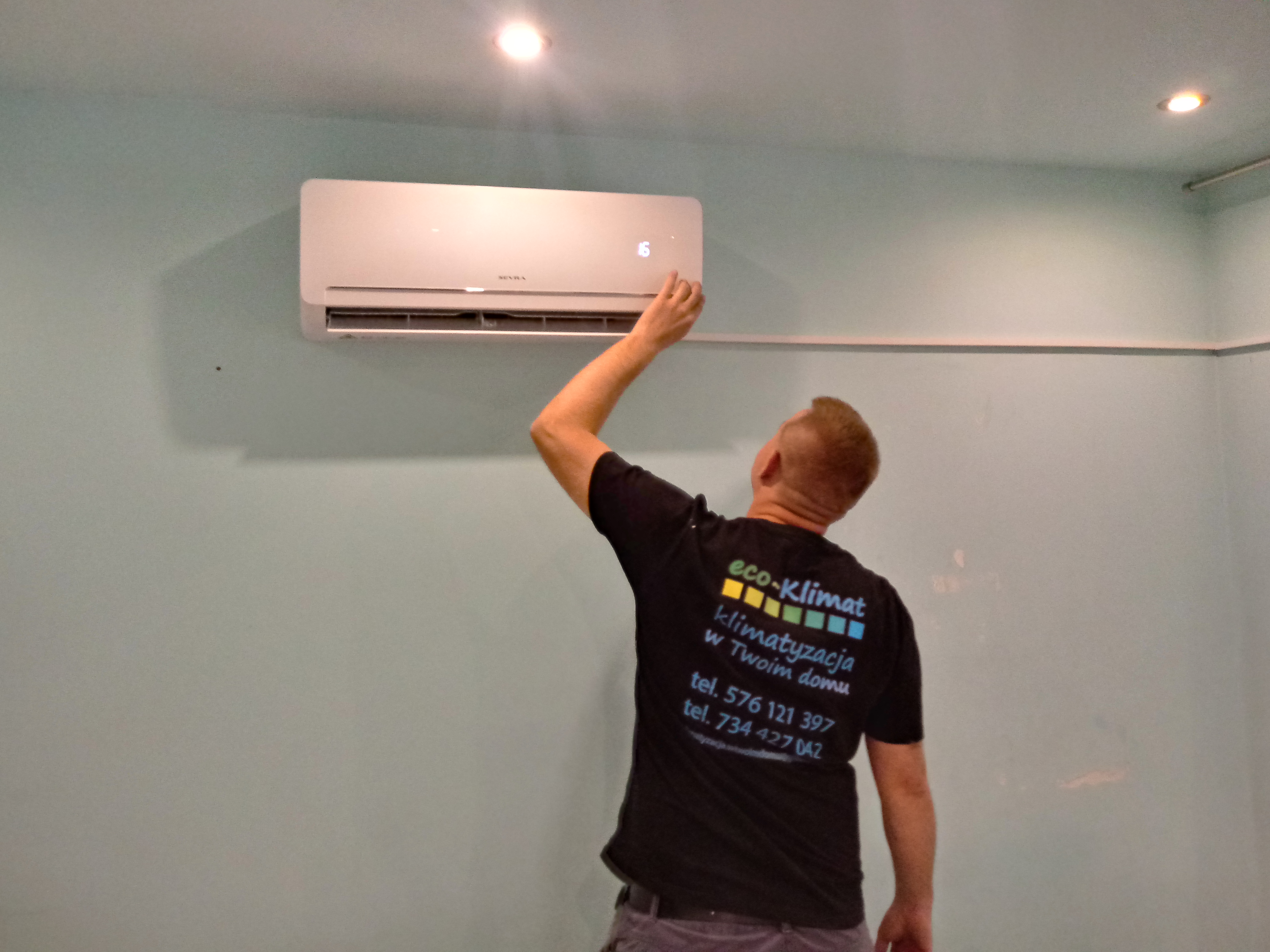 Mężczyzna w czarnej koszulce z logo 'eco-Klimat' ustawia temperaturę na ściennym urządzeniu chłodzącym w jasnym pomieszczeniu.