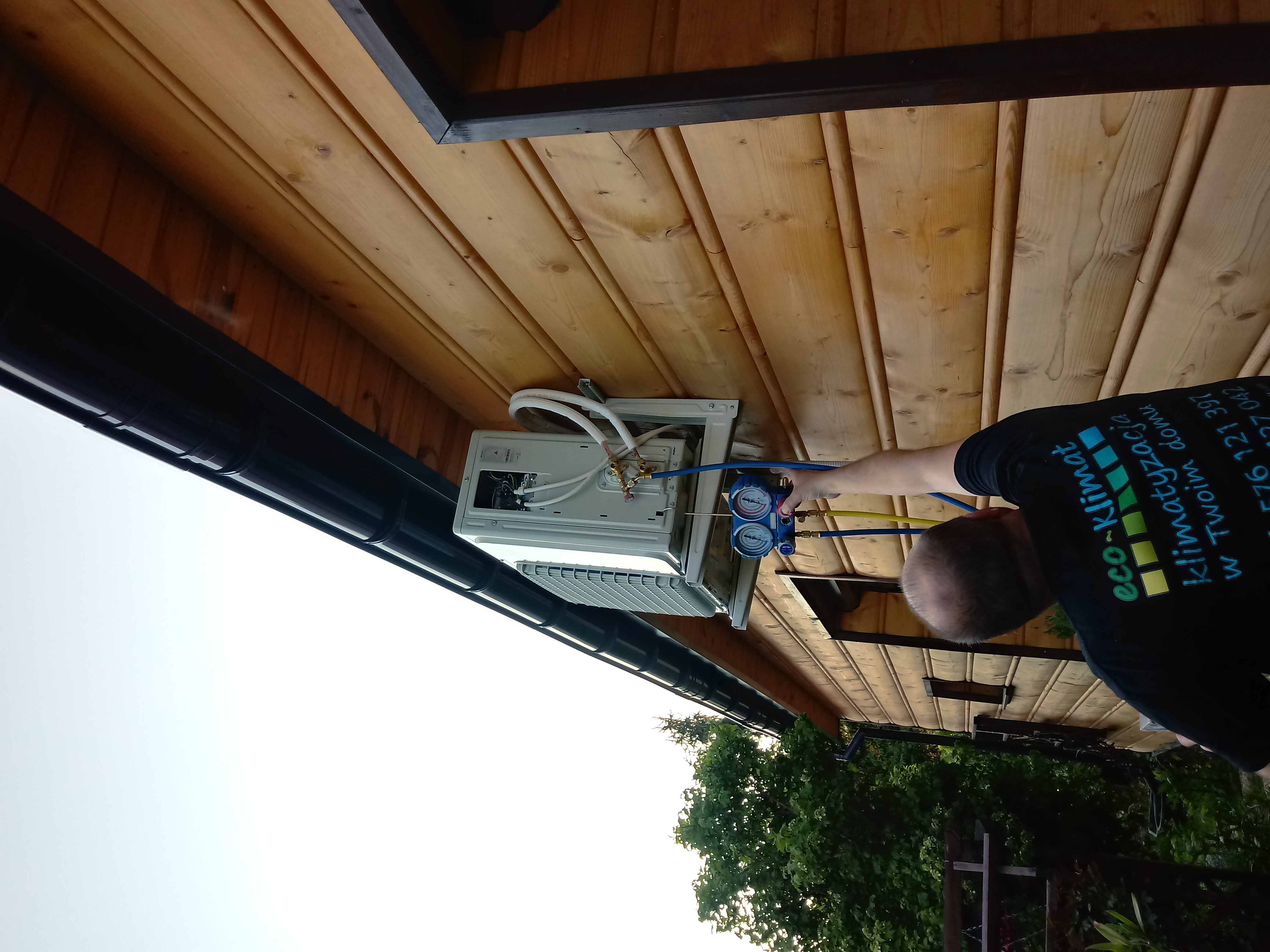 Technik podłącza manometry do zewnętrznej jednostki klimatyzacji zamontowanej pod drewnianym okapem, widoczny fragment koszulki z logo firmy Eco-Klimat.