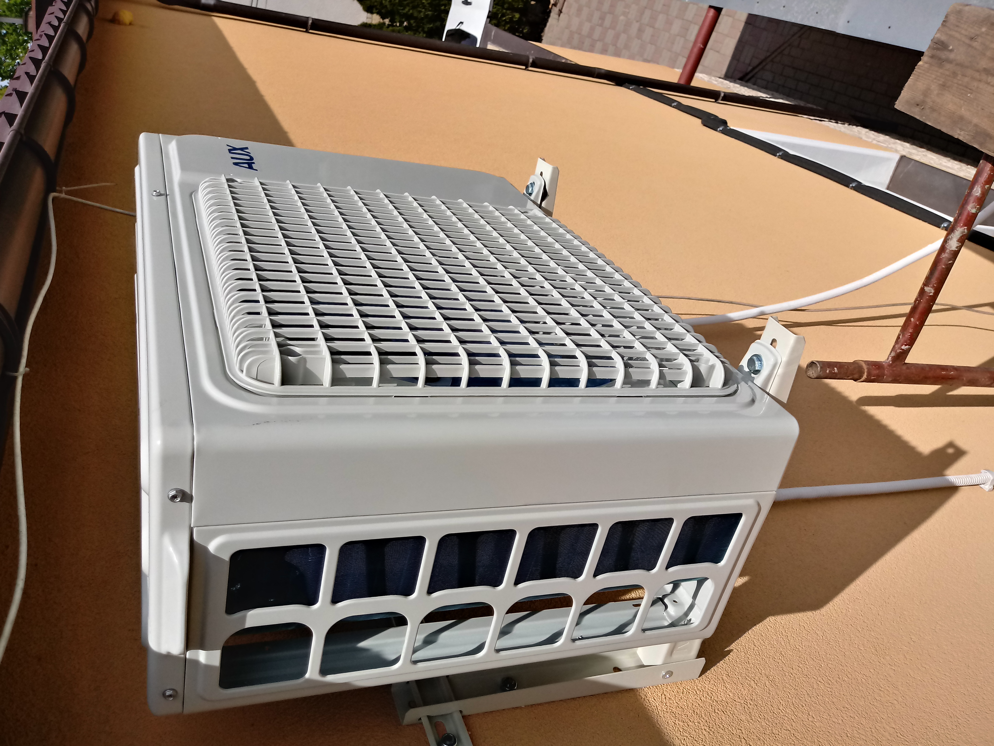 Zewnętrzna jednostka klimatyzatora marki AUX zamontowana na elewacji budynku, widoczna instalacja elektryczna i fragment drabiny.