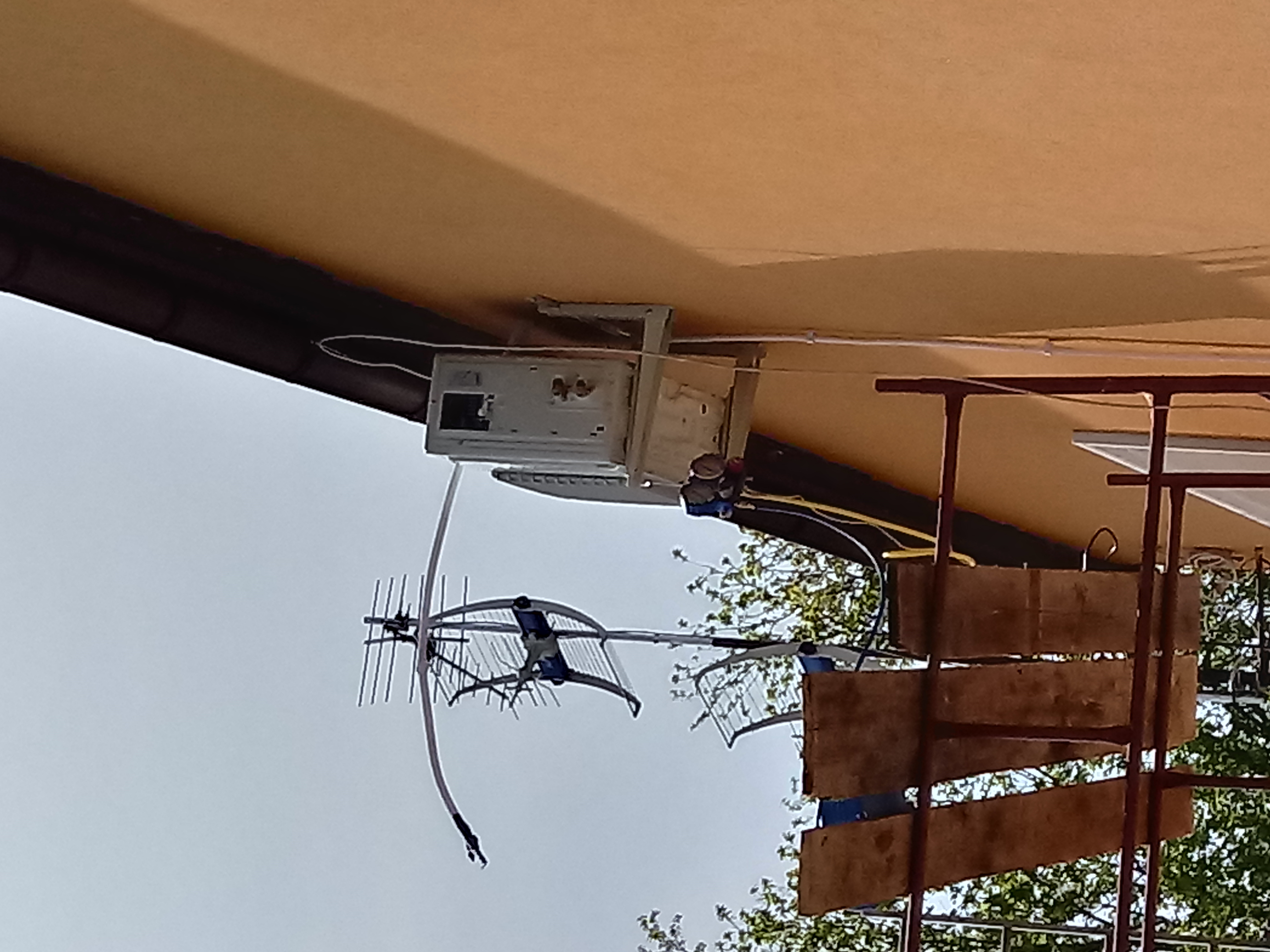 Zewnętrzna jednostka urządzenia przymocowana do elewacji budynku, widoczna instalacja antenowa i fragment konstrukcji metalowej.