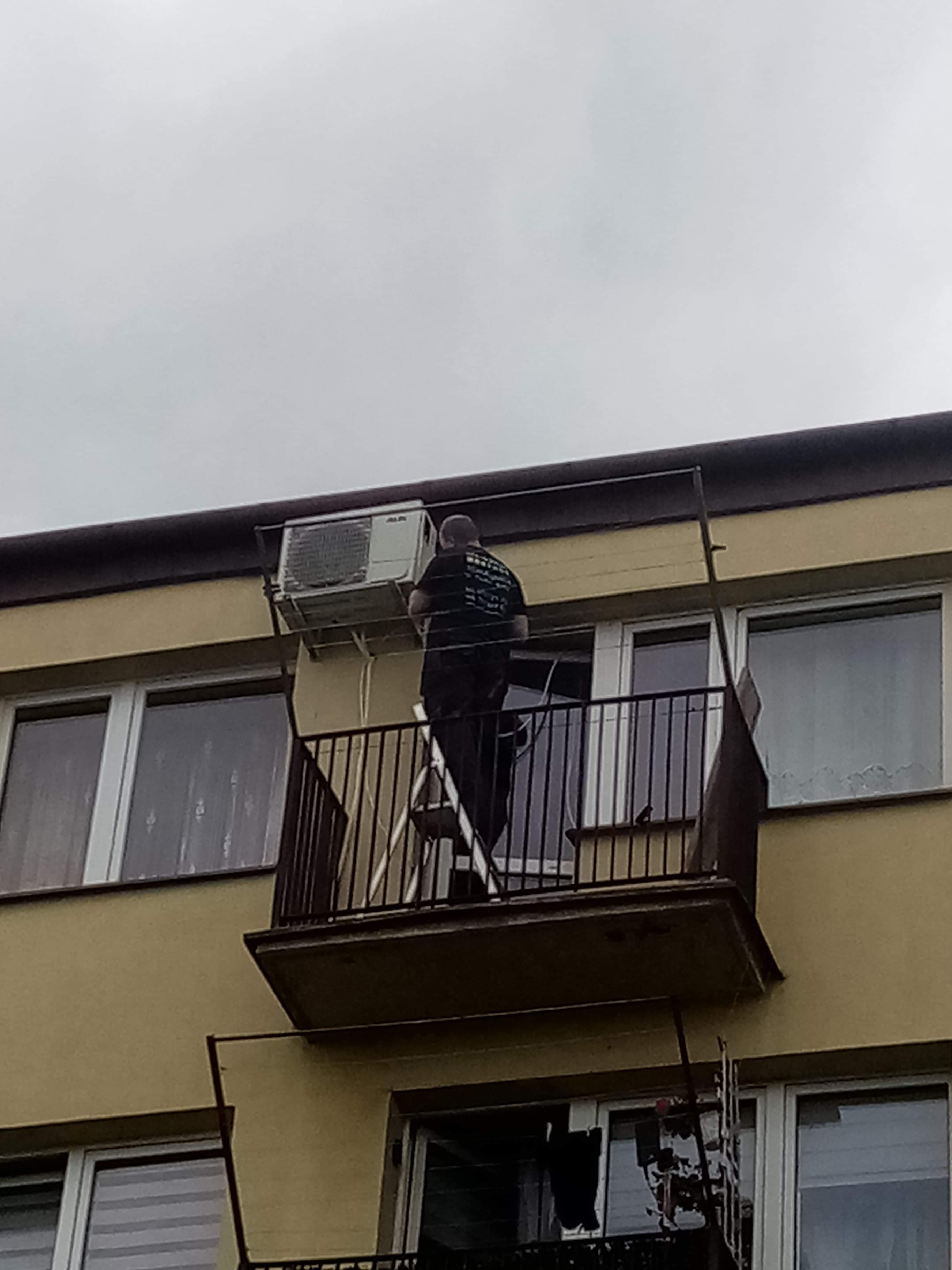 Mężczyzna na drabinie na balkonie montuje jednostkę zewnętrzną klimatyzatora na ścianie budynku mieszkalnego, widok z dołu przy pochmurnym niebie.