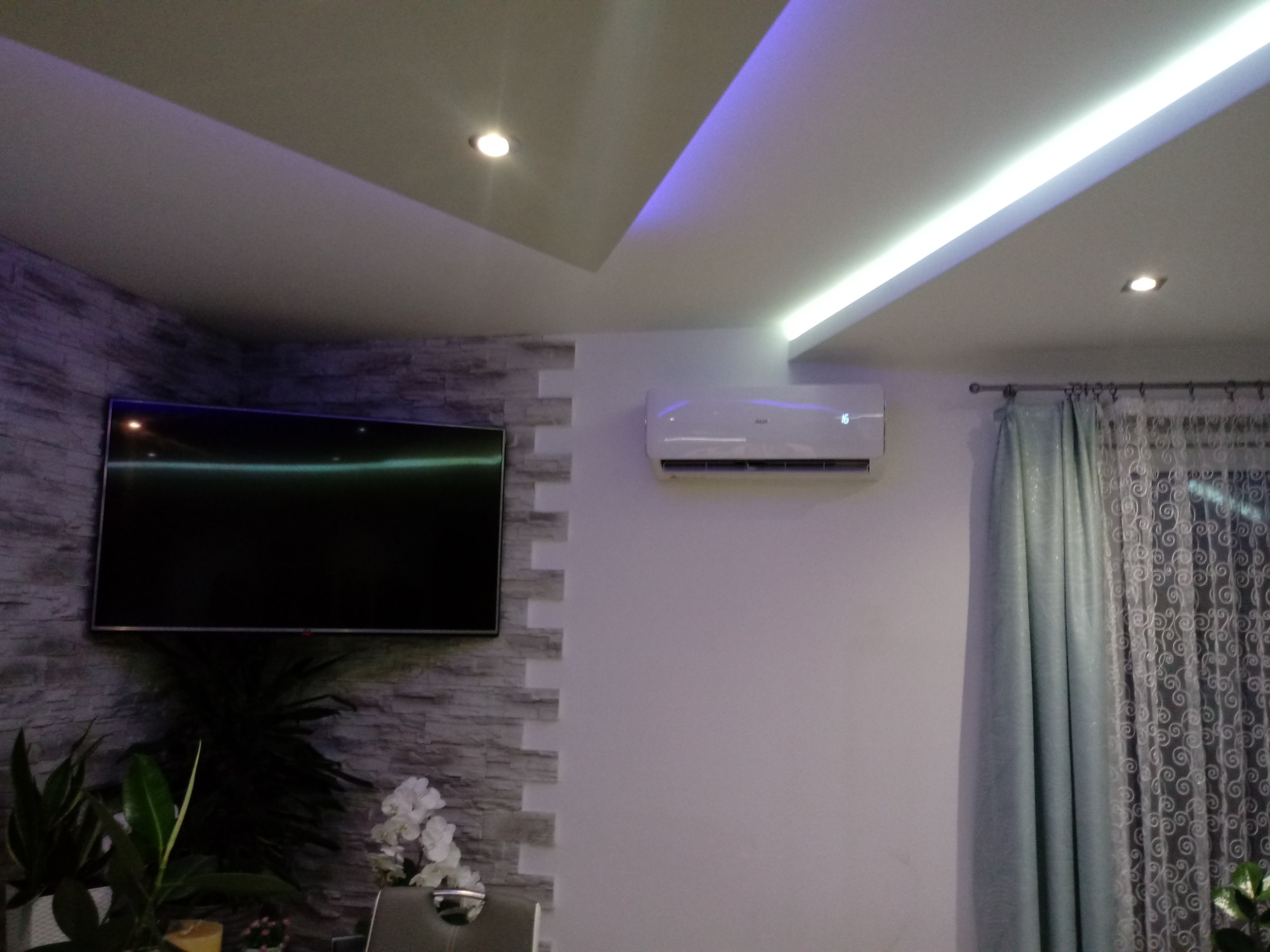 Nowoczesne wnętrze z zamontowanym białym klimatyzatorem ściennym, oświetleniem LED i telewizorem na ścianie z dekoracyjną okładziną imitującą kamień, uzupełnione roślinami doniczkowymi i zasłonami.
