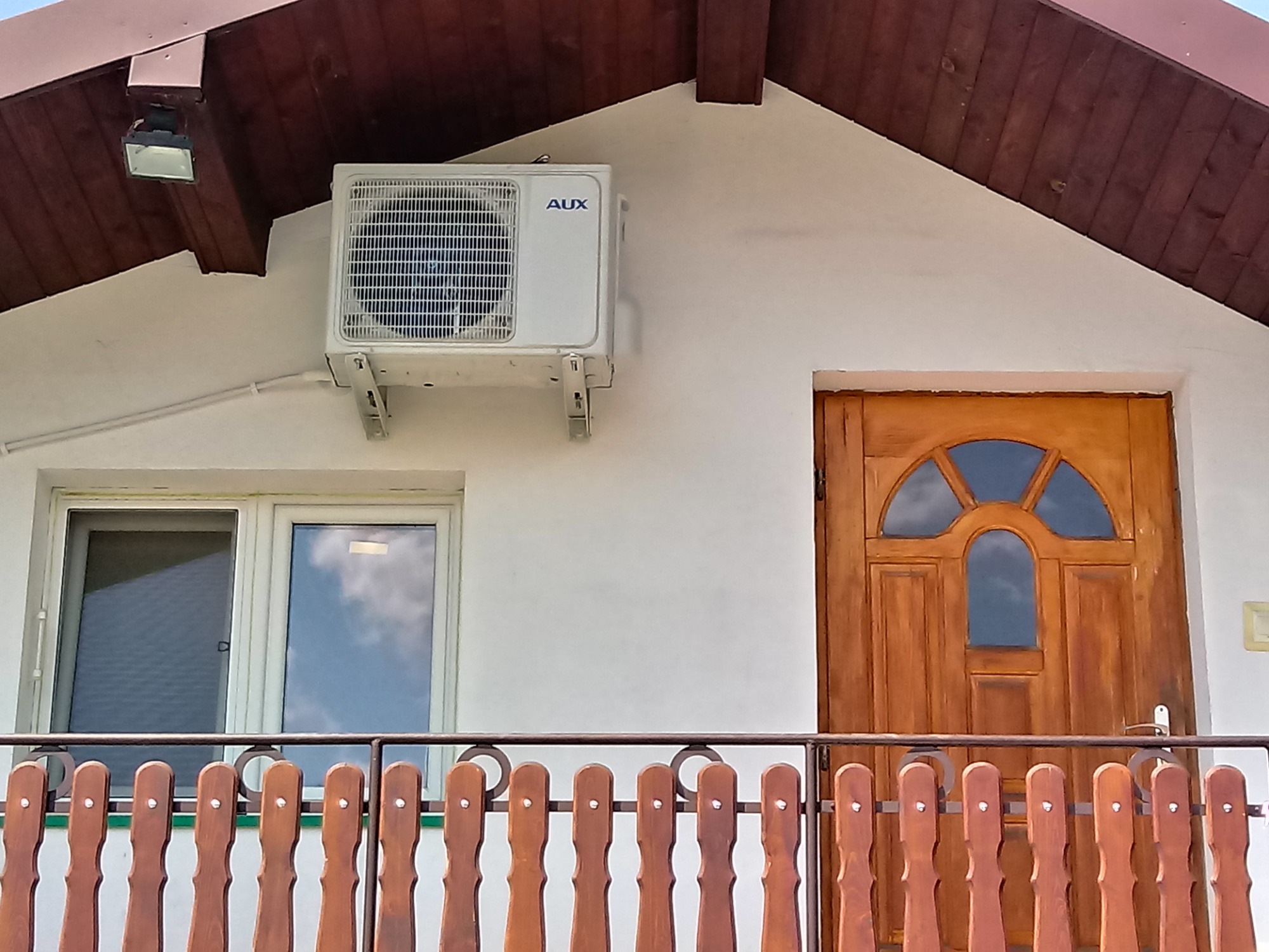 Biały klimatyzator marki AUX zamontowany na elewacji domu obok okna i drewnianych drzwi wejściowych, widok z balkonu z drewnianą balustradą.