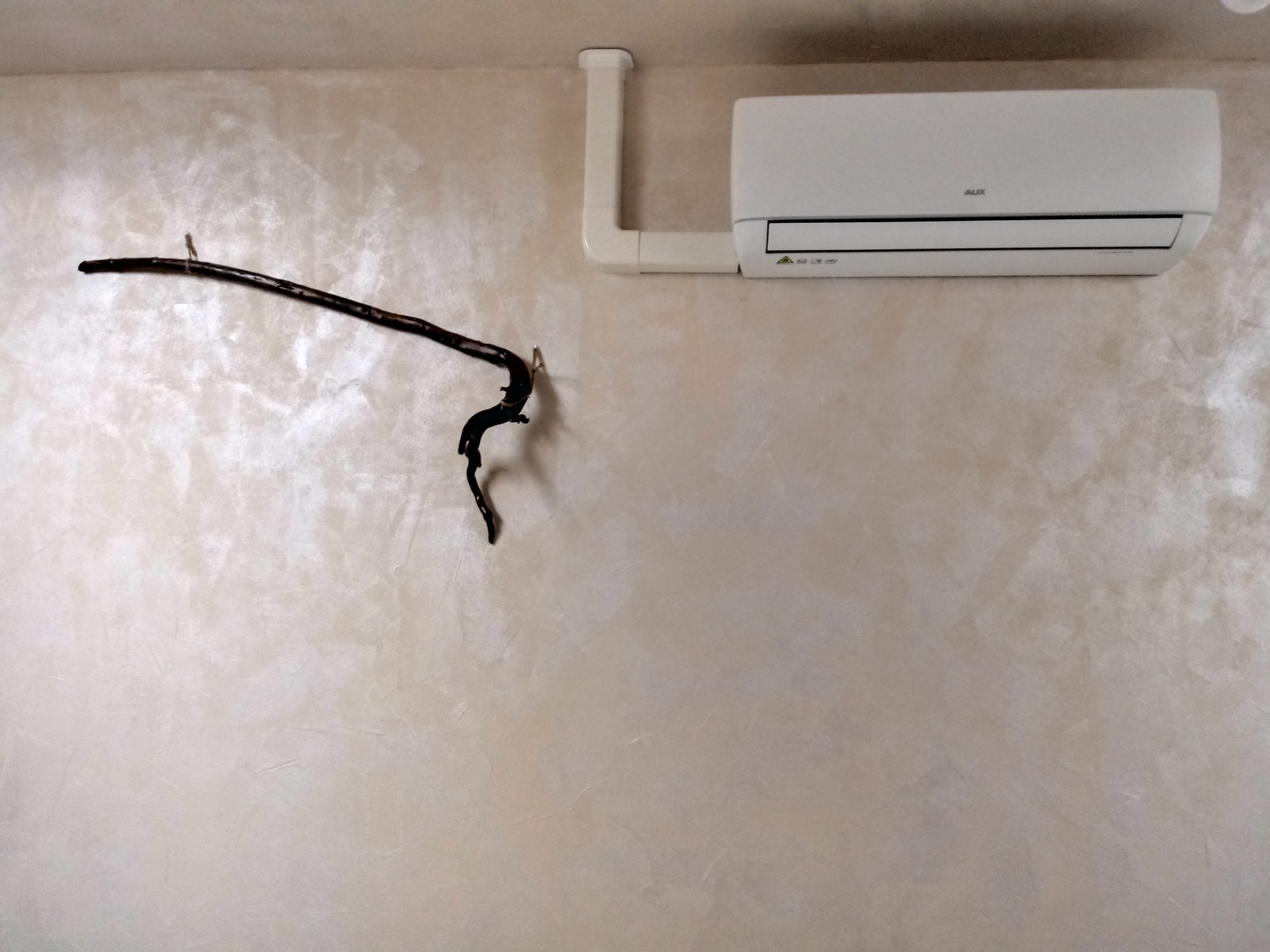 Biały klimatyzator marki AUX zamontowany na ścianie o dekoracyjnej fakturze, z widoczną rurą odprowadzającą.