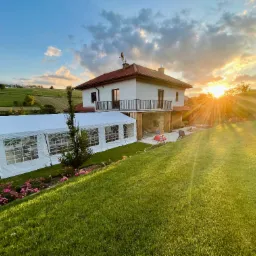 Biały namiot bankietowy z oknami, ustawiony na trawie przed domem jednorodzinnym w promieniach zachodzącego słońca.