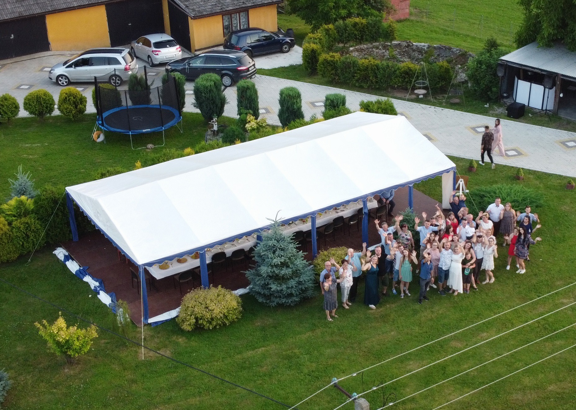 Duży biały namiot z niebieskimi akcentami na drewnianym podeście, ustawiony na trawniku, z grupą ludzi machających w tle, widok z góry.