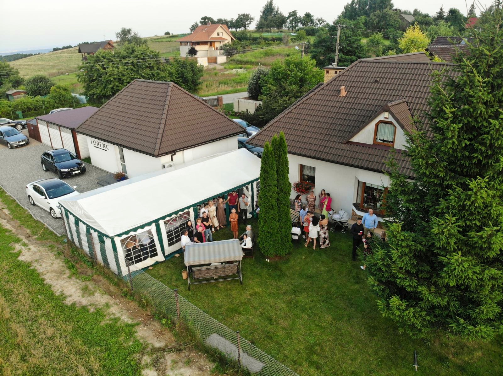 Widok z góry na biały namiot z zielonymi akcentami, ustawiony na trawniku przed budynkiem z grupą ludzi wewnątrz i na zewnątrz, sugerujący uroczystość plenerową.