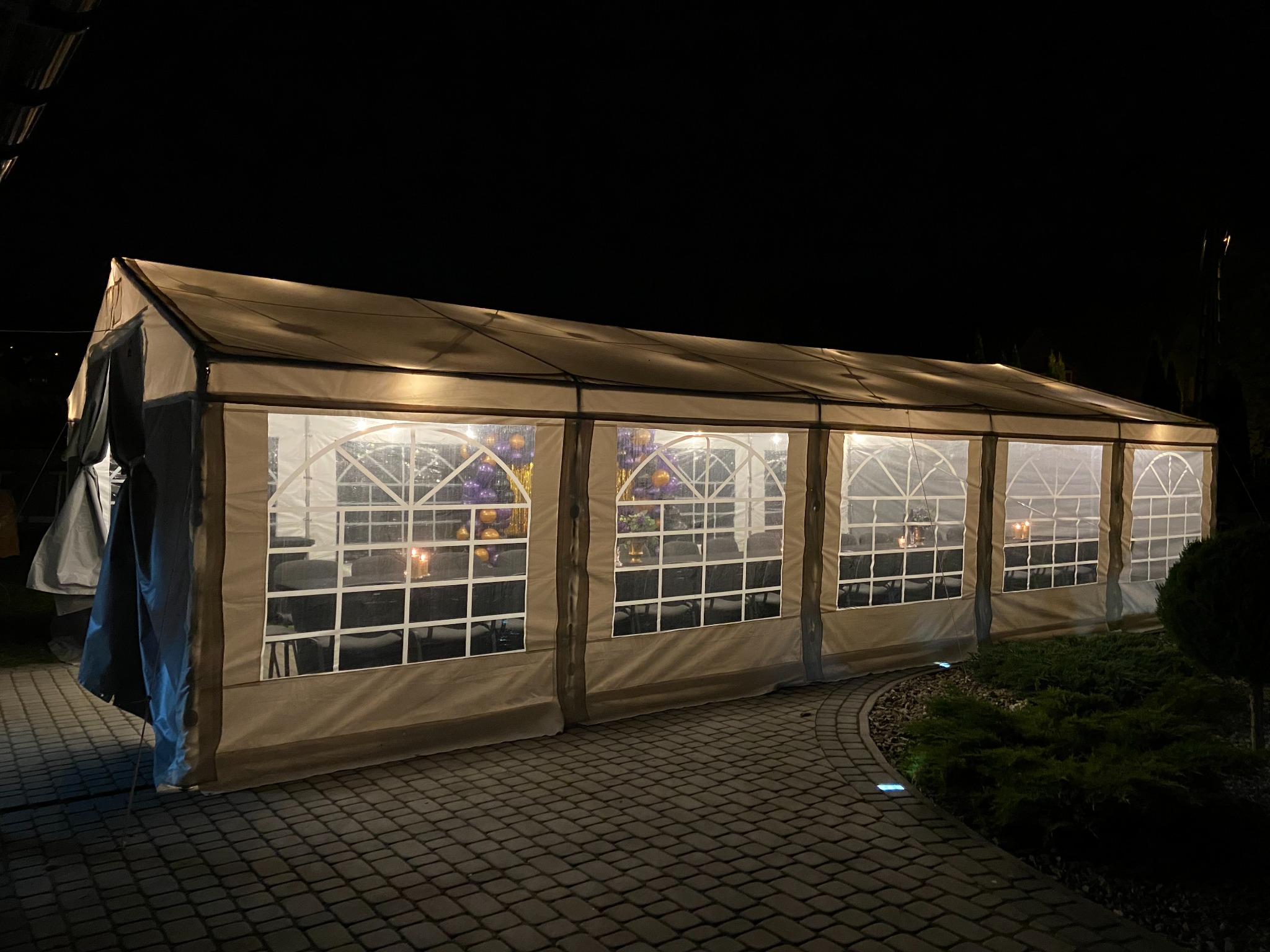 Długi, oświetlony namiot bankietowy z przezroczystymi ścianami ustawiony na brukowanym podłożu, widoczne stoły i krzesła wewnątrz, dekoracje kwiatowe i świece na stołach, ciemne tło nocne.