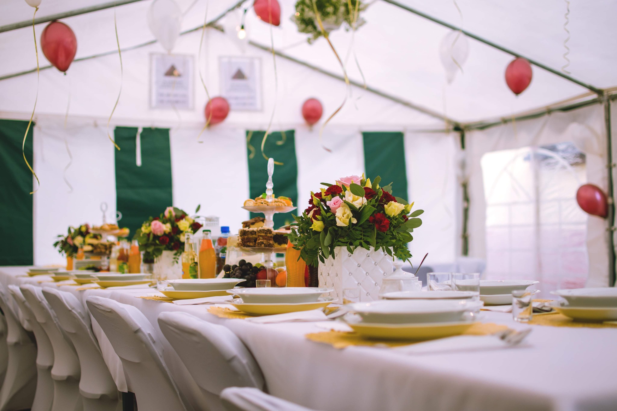Uroczysty stół w namiocie, nakryty białym obrusem, z kwiatowymi dekoracjami w białych wazonach, talerzami z żółtym rantem, napojami w butelkach i balonami wiszącymi pod sufitem.