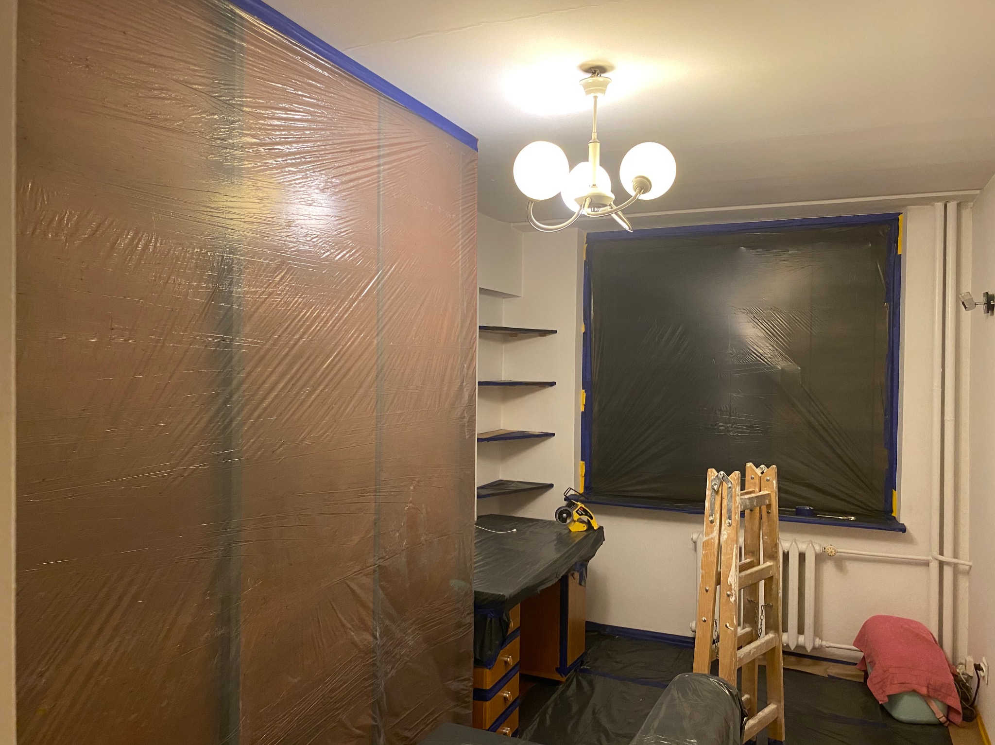 Pokój przygotowany do malowania: meble i okna zabezpieczone folią, ściany oklejone taśmą malarską. Widoczna drabina i oświetlenie sufitowe.