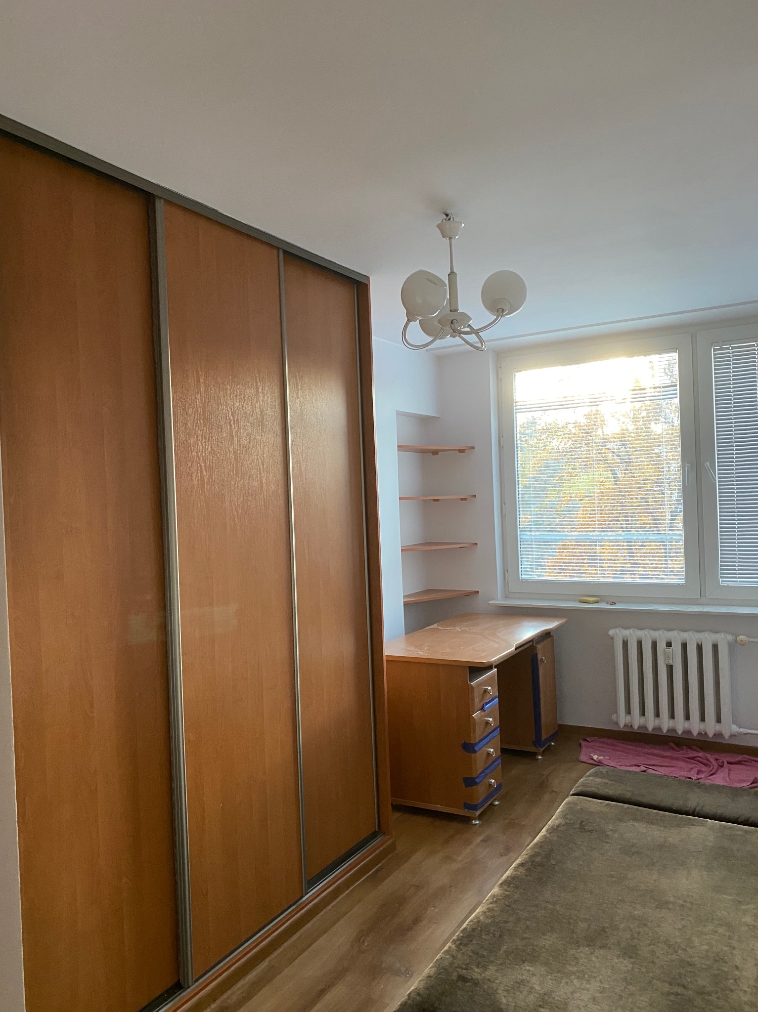 Pokój z dużą szafą przesuwną z drewnianymi frontami, biurkiem przy oknie i półkami na ścianie. Widoczne panele podłogowe i grzejnik. Oświetlenie sufitowe.