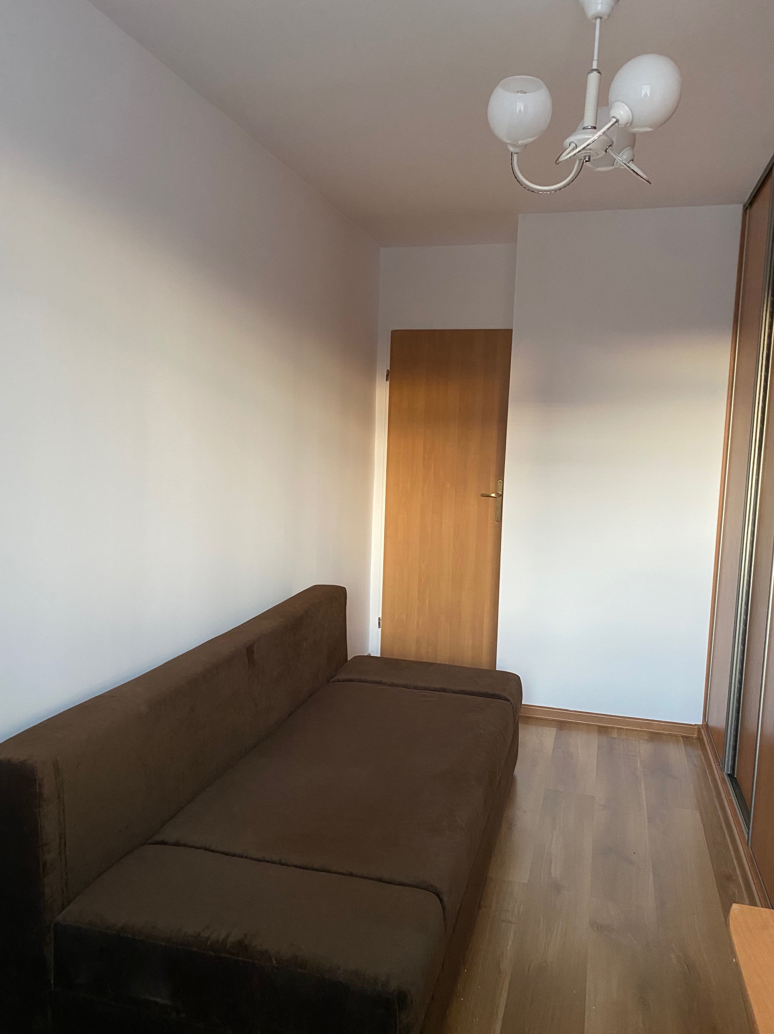 Ustawiony brązowy narożnik w pokoju z drewnianą podłogą i drzwiami. Widoczna biała ściana i sufit z lampą. Ujęcie z niskiego kąta, podkreślające przestrzeń.