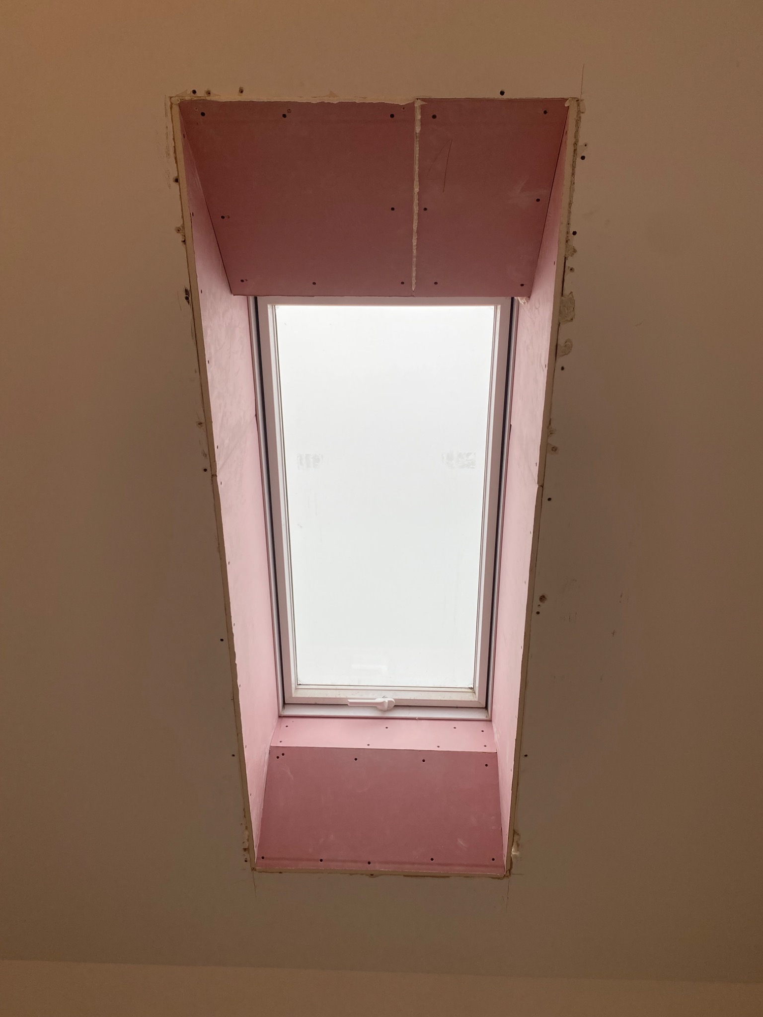 Montaż płyty GK wokół okna dachowego, widok od dołu. Różowa płyta karton-gips, widoczne ślady po wkrętach. Białe okno dachowe w skośnym dachu. Etap prac wykończeniowych.