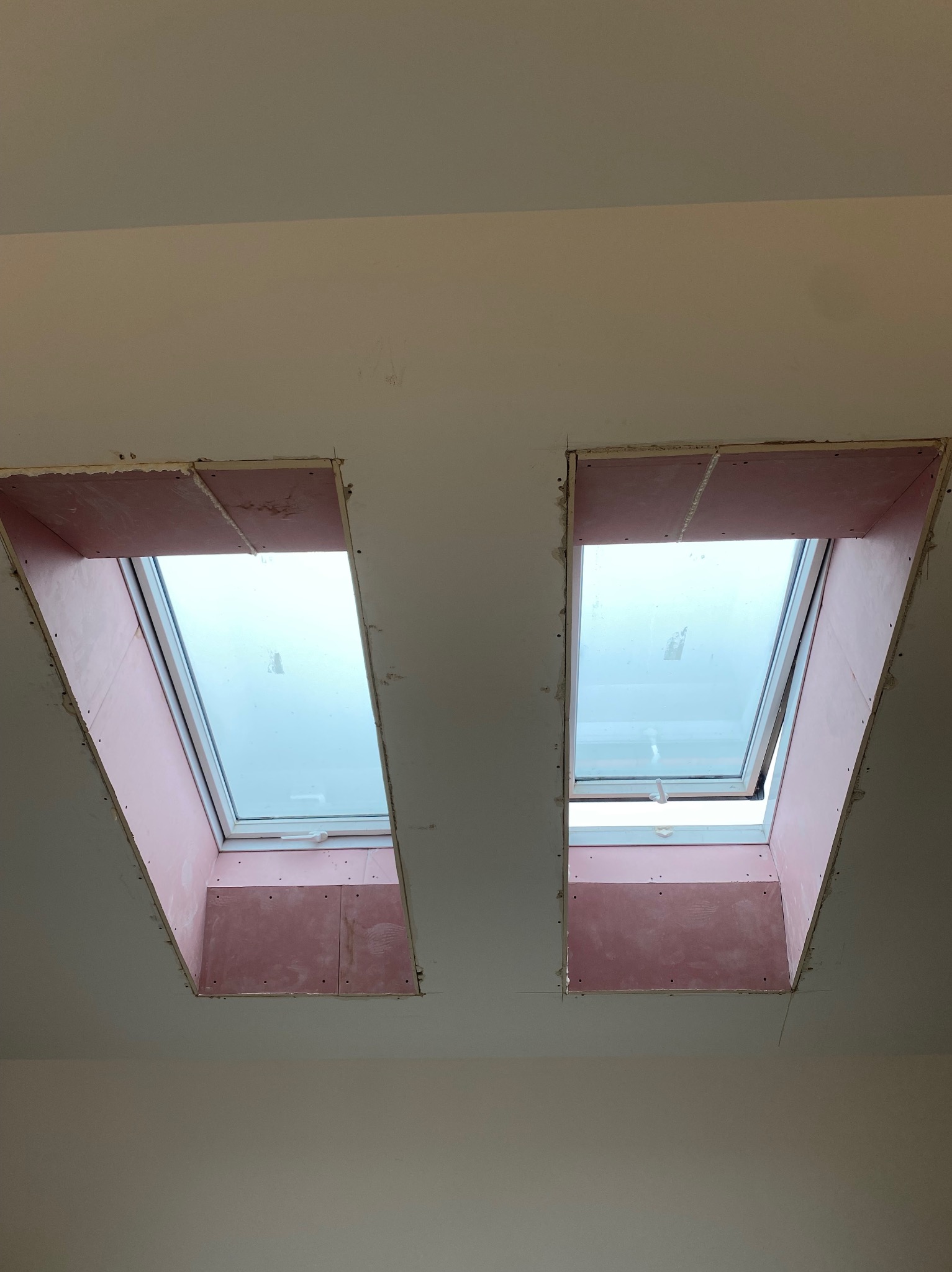 Dwa okna dachowe w trakcie montażu w suficie, obudowane płytami kartonowo-gipsowymi w kolorze różowym, widoczne łączenia i śruby. Surowy stan wykończenia.