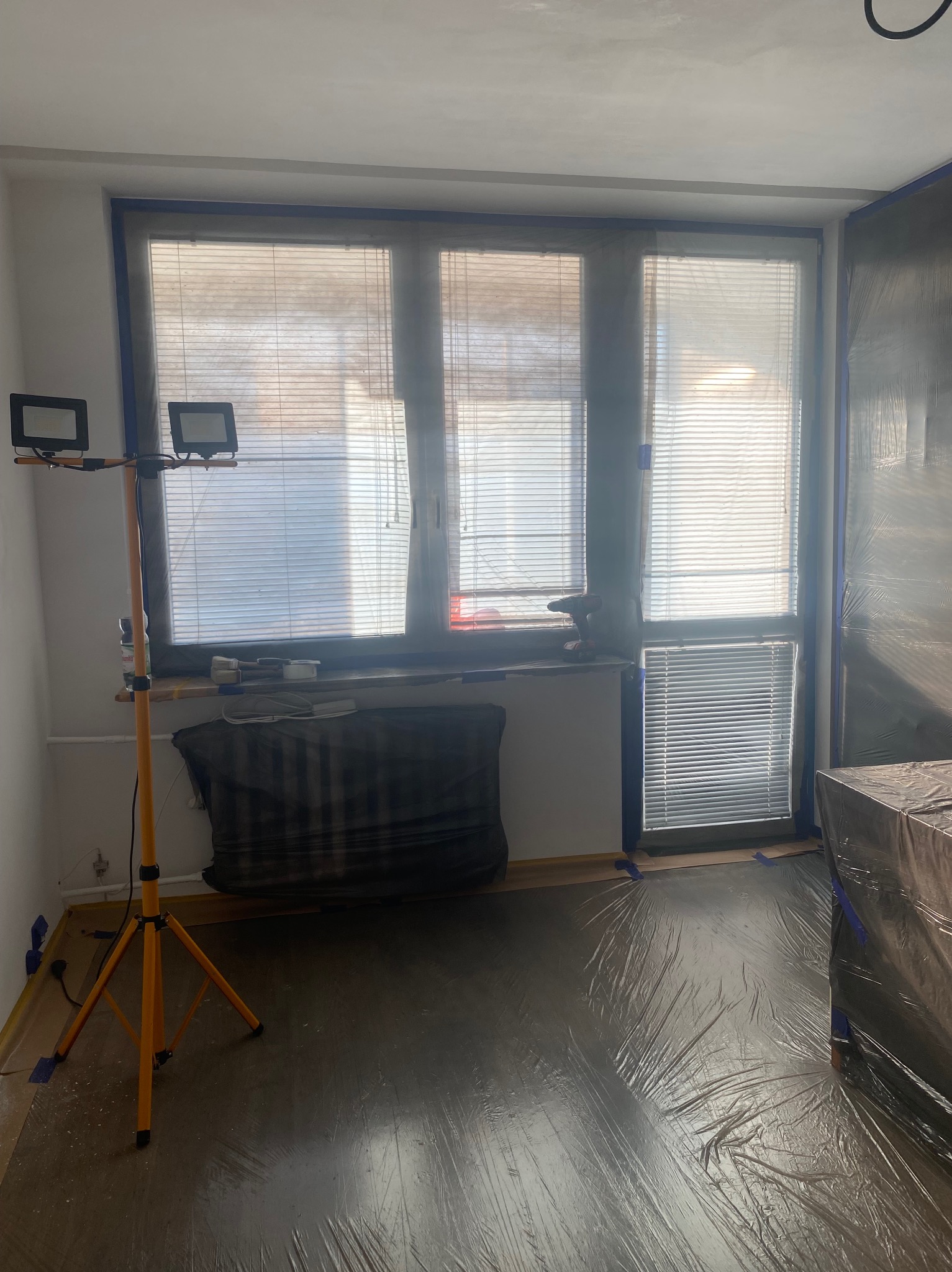 Pokój przygotowany do malowania: okna i framugi zabezpieczone taśmą, podłoga folią ochronną, lampa budowlana. Widoczne narzędzia malarskie na parapecie.