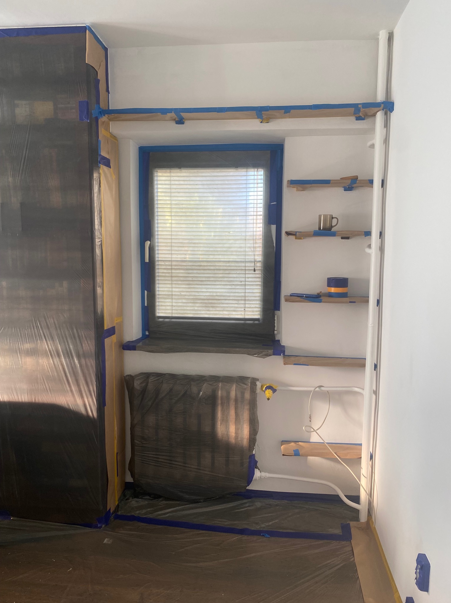 Pokój przygotowany do malowania: okno, grzejnik i meble zabezpieczone folią i taśmą malarską. Półki z kubkiem i dekoracją. Widoczne rury grzewcze.