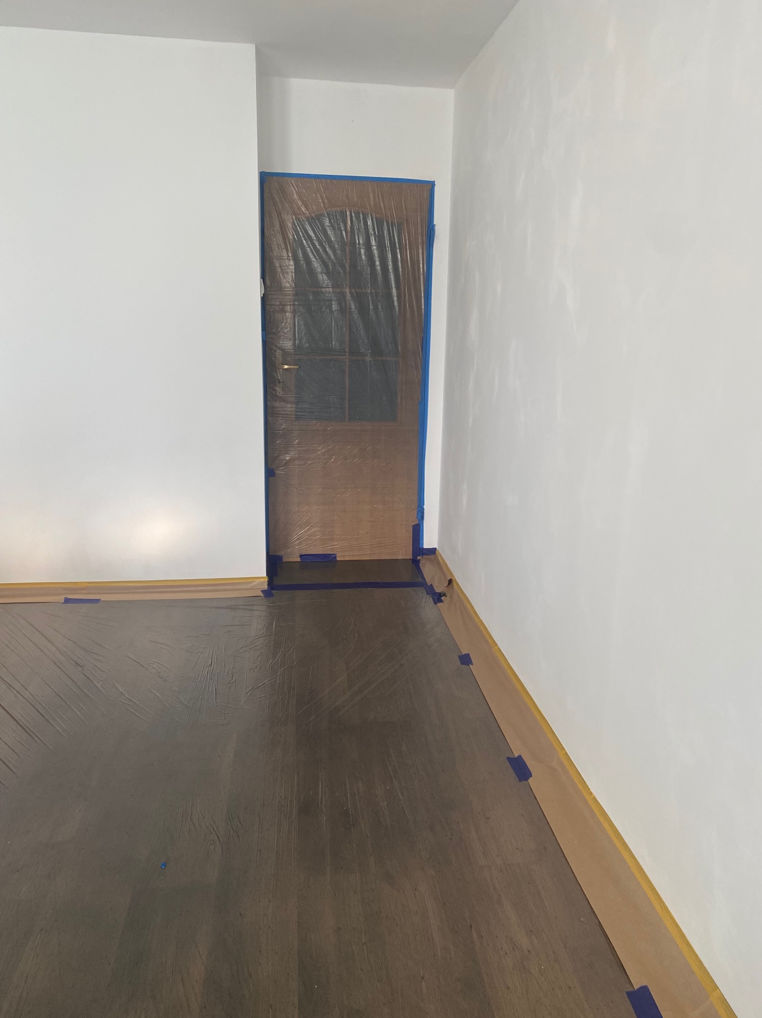 Przygotowanie do malowania: zabezpieczona folią i taśmą drzwi i podłoga z parkietu w pokoju z białymi ścianami. Widoczne detale zabezpieczeń.