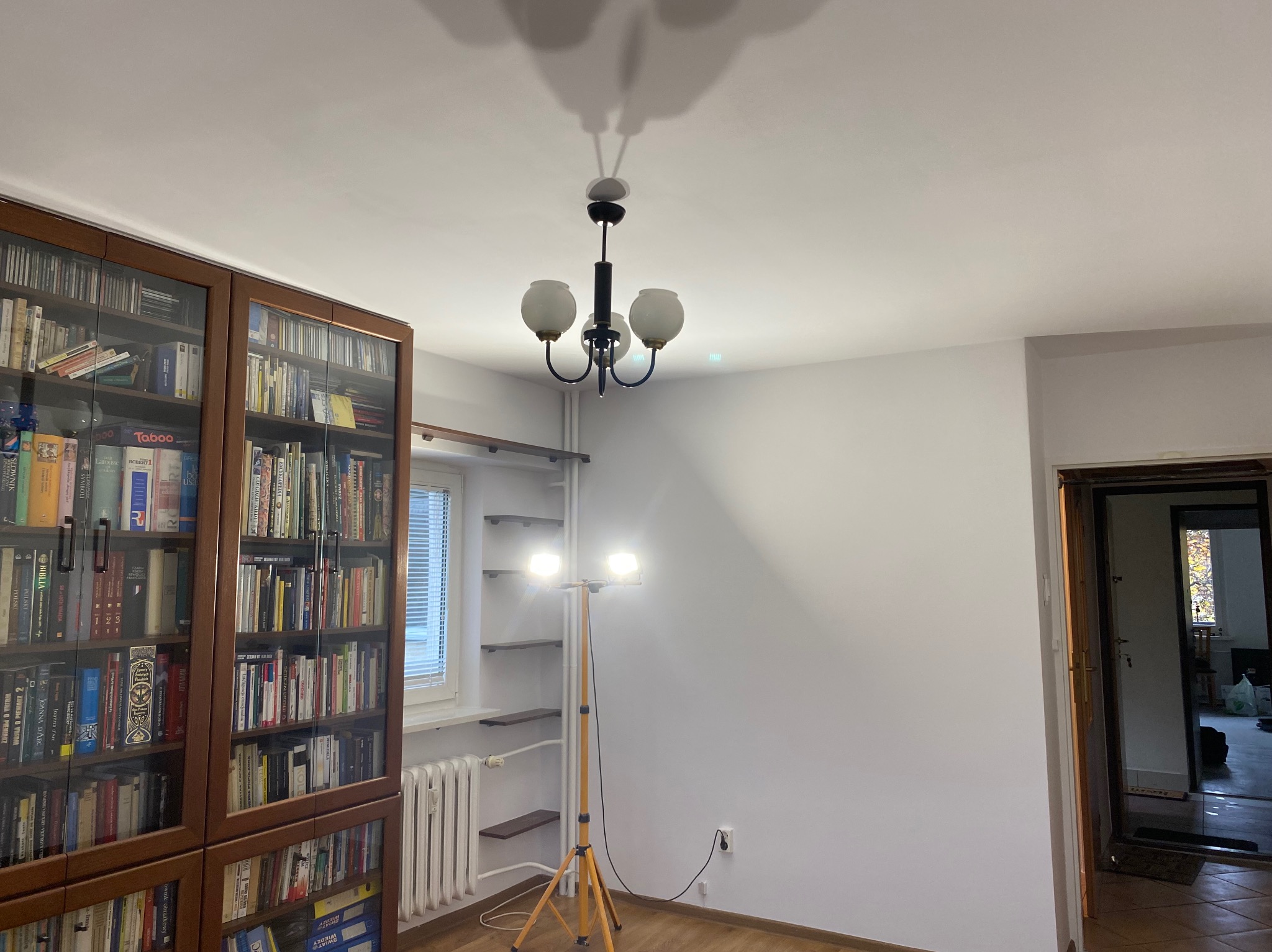 Pokój przed remontem: regał z książkami, lampa sufitowa, grzejnik, lampa na statywie. Widoczne ślady użytkowania, przygotowanie do prac wykończeniowych.