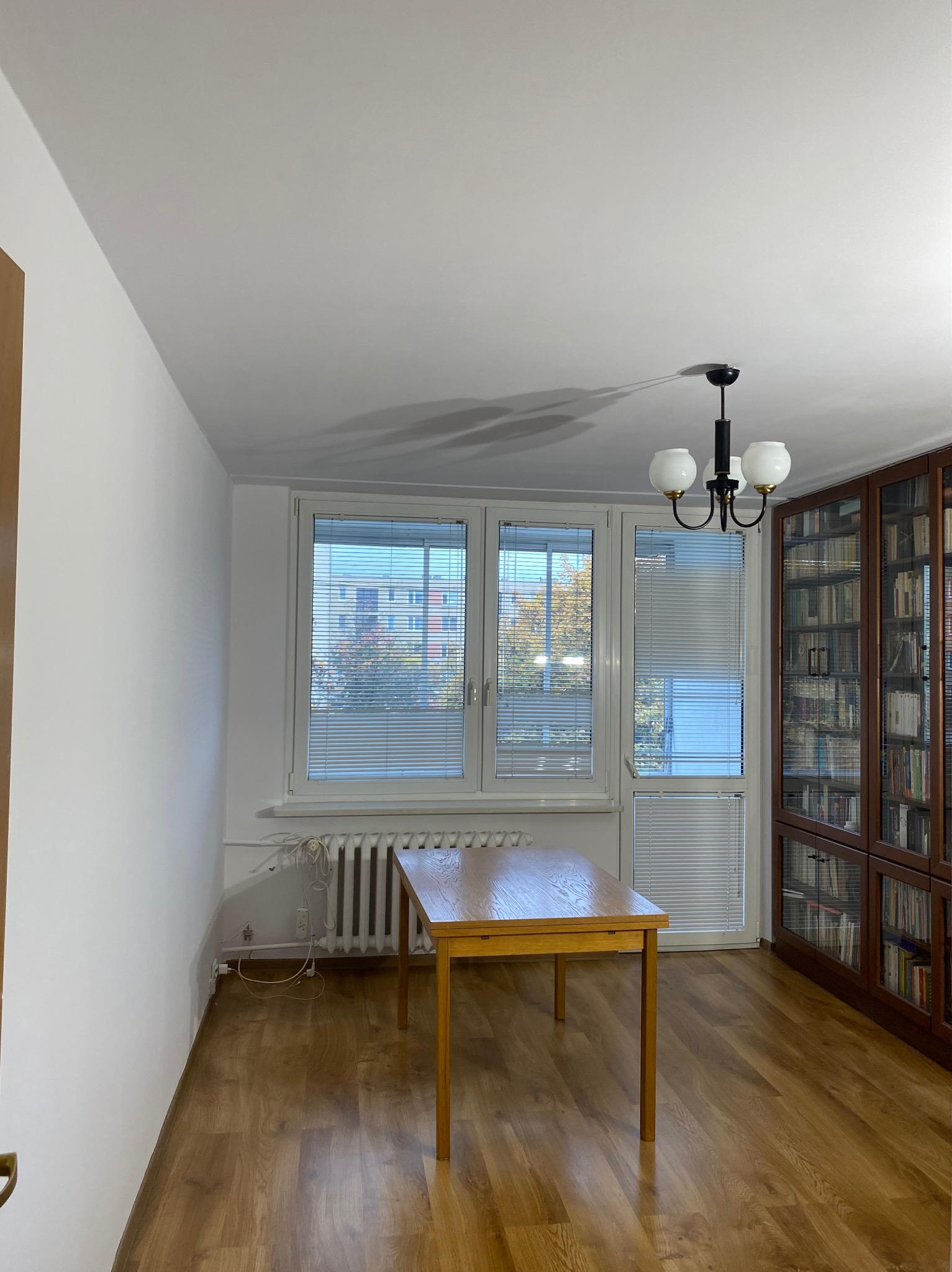 Jasne wnętrze pokoju po odświeżeniu: białe ściany, drewniana podłoga, stół, okno z żaluzjami i regał z książkami. Widoczne ślady po remoncie.
