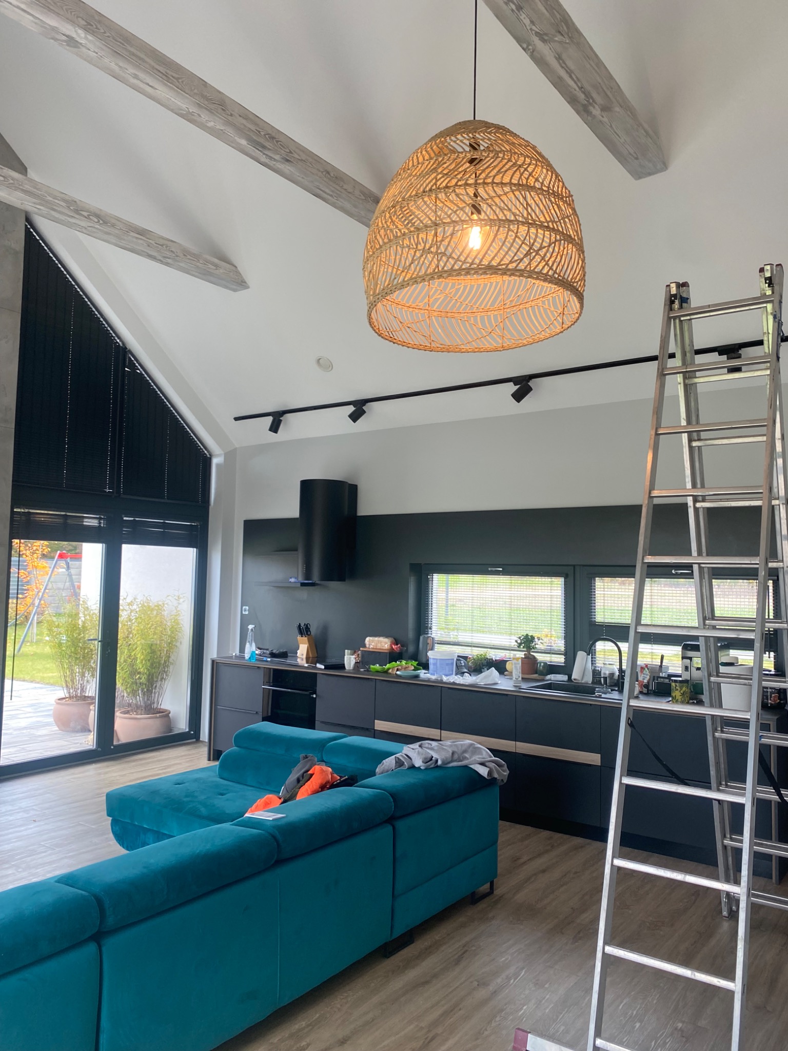 Nowoczesne wnętrze z turkusową sofą, drewnianymi belkami stropowymi i rattanową lampą. Aluminiowa drabina oparta o ścianę. Widok na czarną zabudowę kuchenną.