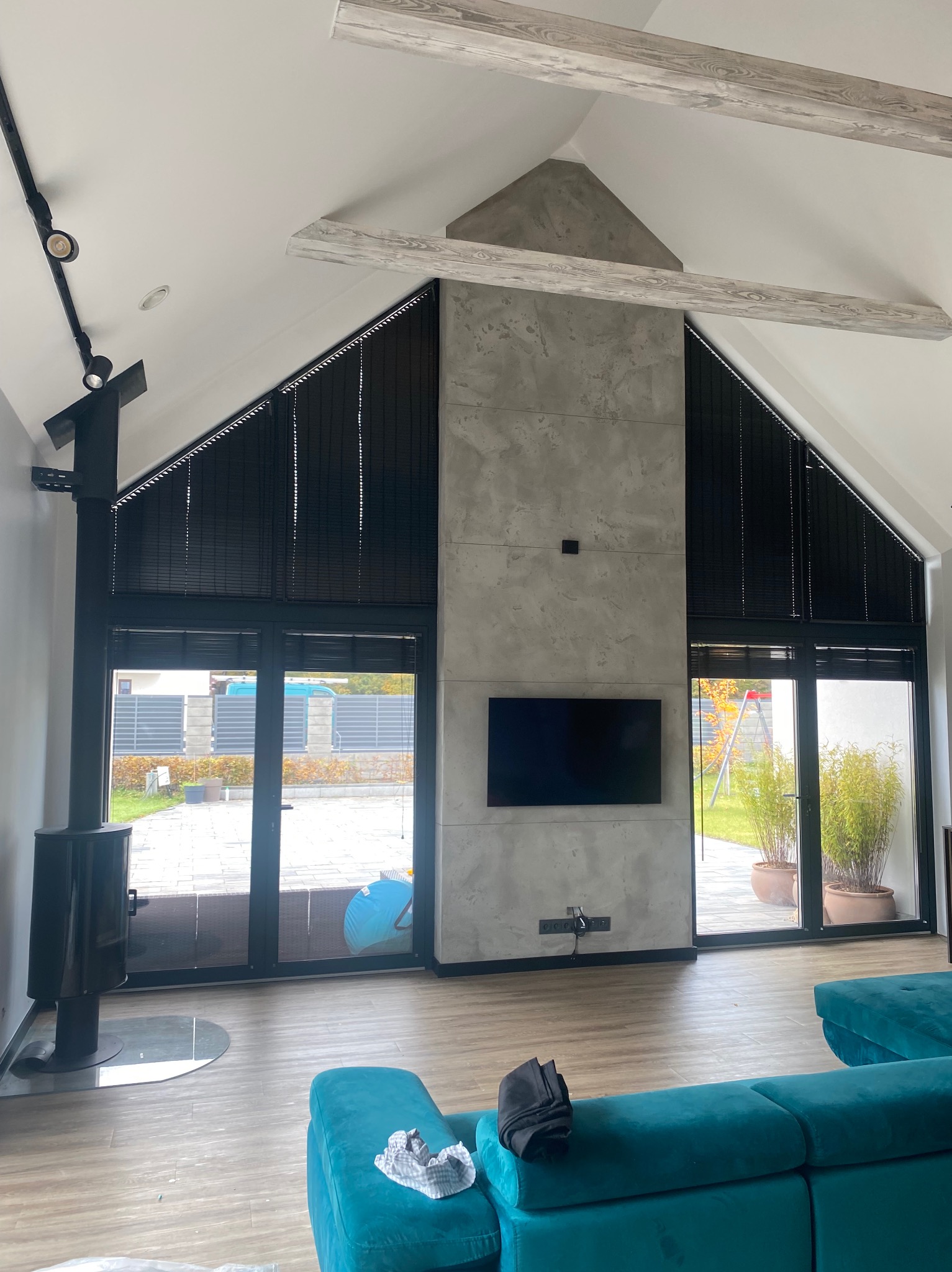 Nowoczesny salon z wysokim sufitem, betonową ścianą i dużymi oknami z żaluzjami. Widoczny kominek, turkusowa sofa i drewniane belki stropowe.