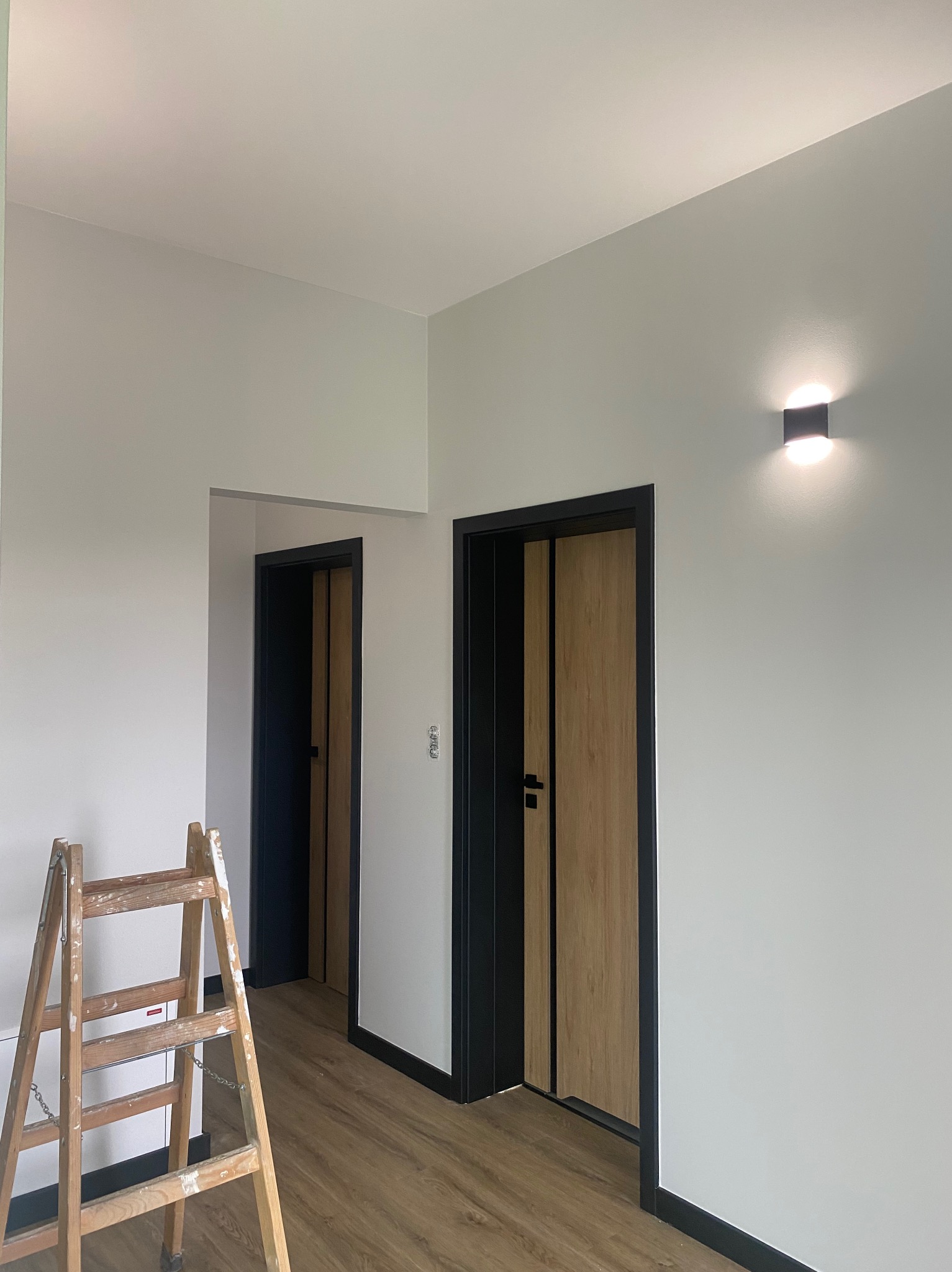 Wnętrze po remoncie: świeżo pomalowane ściany w odcieniach szarości, dwoje drzwi z czarnymi ościeżnicami i drewnianymi skrzydłami, drewniana podłoga, drabina malarska.
