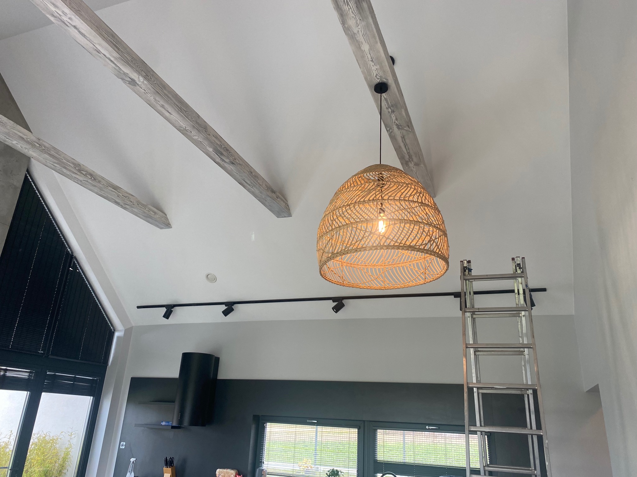 Wnętrze z drewnianymi belkami stropowymi, oświetleniem szynowym i wiklinową lampą. Aluminiowa drabina oparta o ścianę. Widok na okno i fragment wyposażenia kuchni.