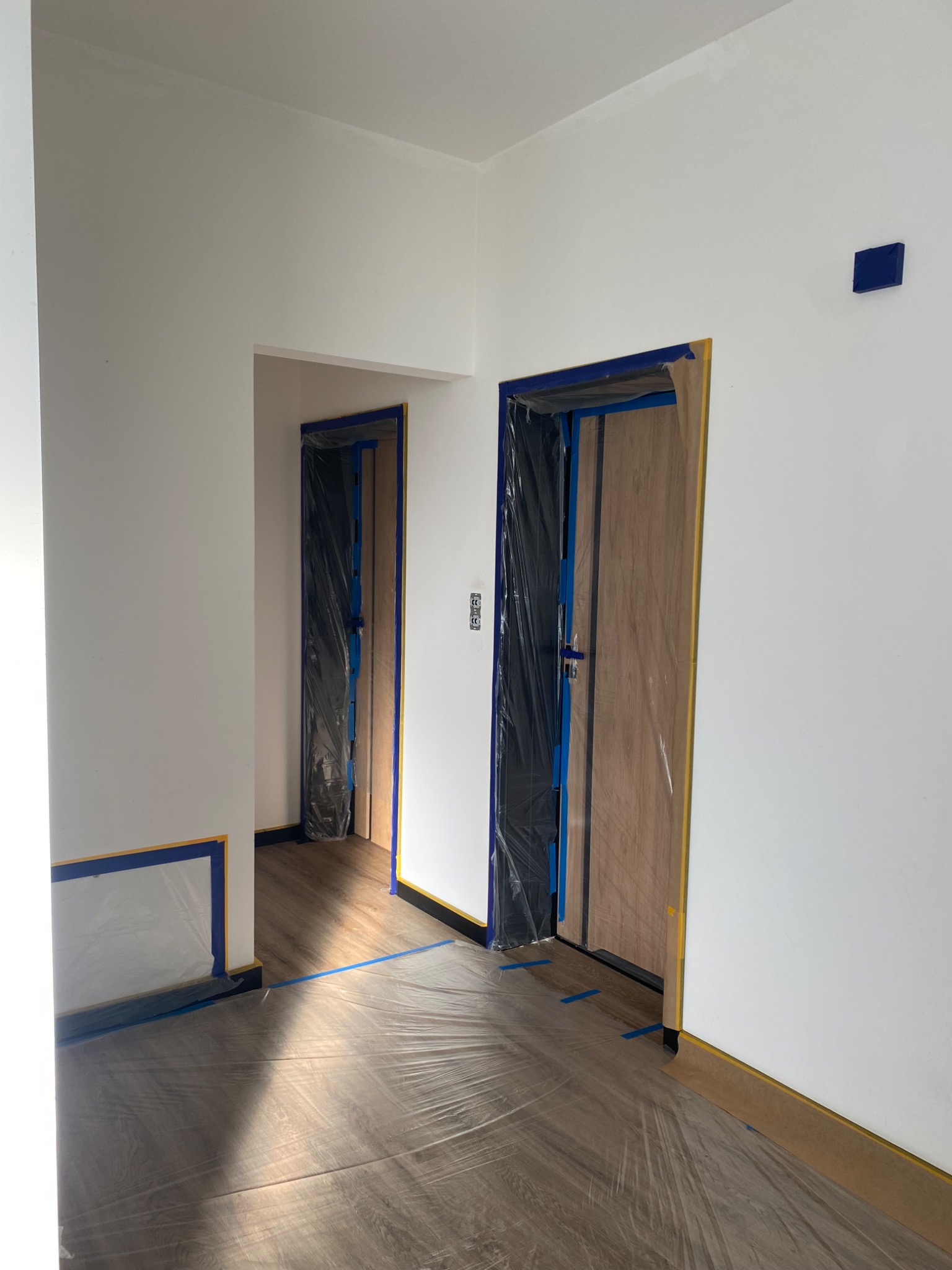 Przygotowanie do malowania: zabezpieczone folią i taśmą drzwi, podłoga i ściany w jasnym wnętrzu. Widoczne detale wykończeniowe i fragment podłogi.