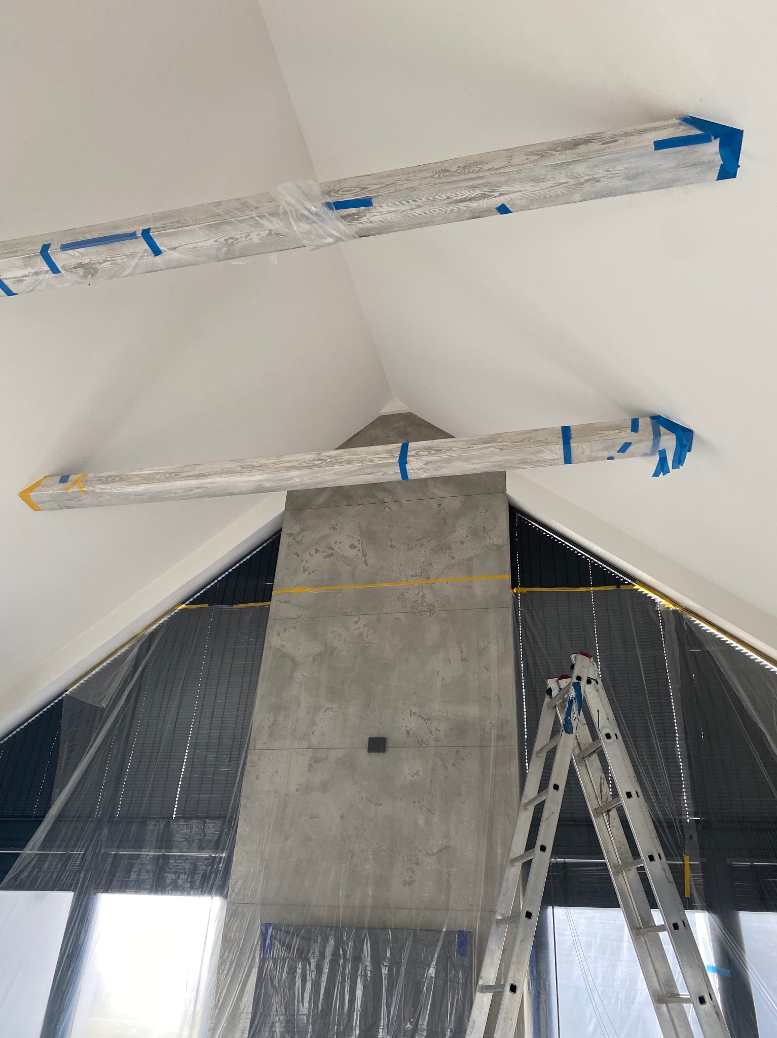 Skośny sufit z drewnianymi belkami zabezpieczonymi folią i taśmą malarską, betonowa ściana, drabina. Przygotowania do prac wykończeniowych.