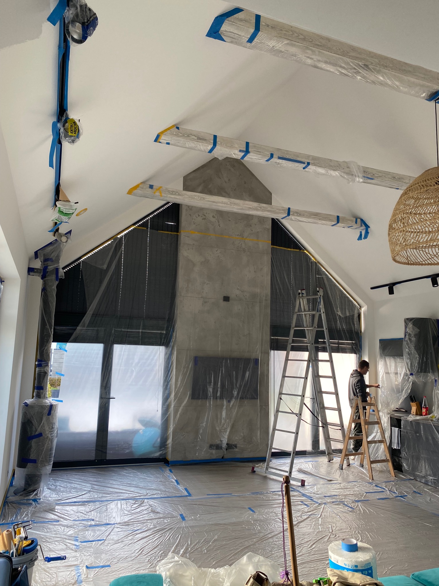 Przygotowanie do prac malarskich w przestronnym salonie z wysokim sufitem. Folia ochronna zabezpiecza podłogę i meble. Widoczna drabina i osoba malująca ścianę.