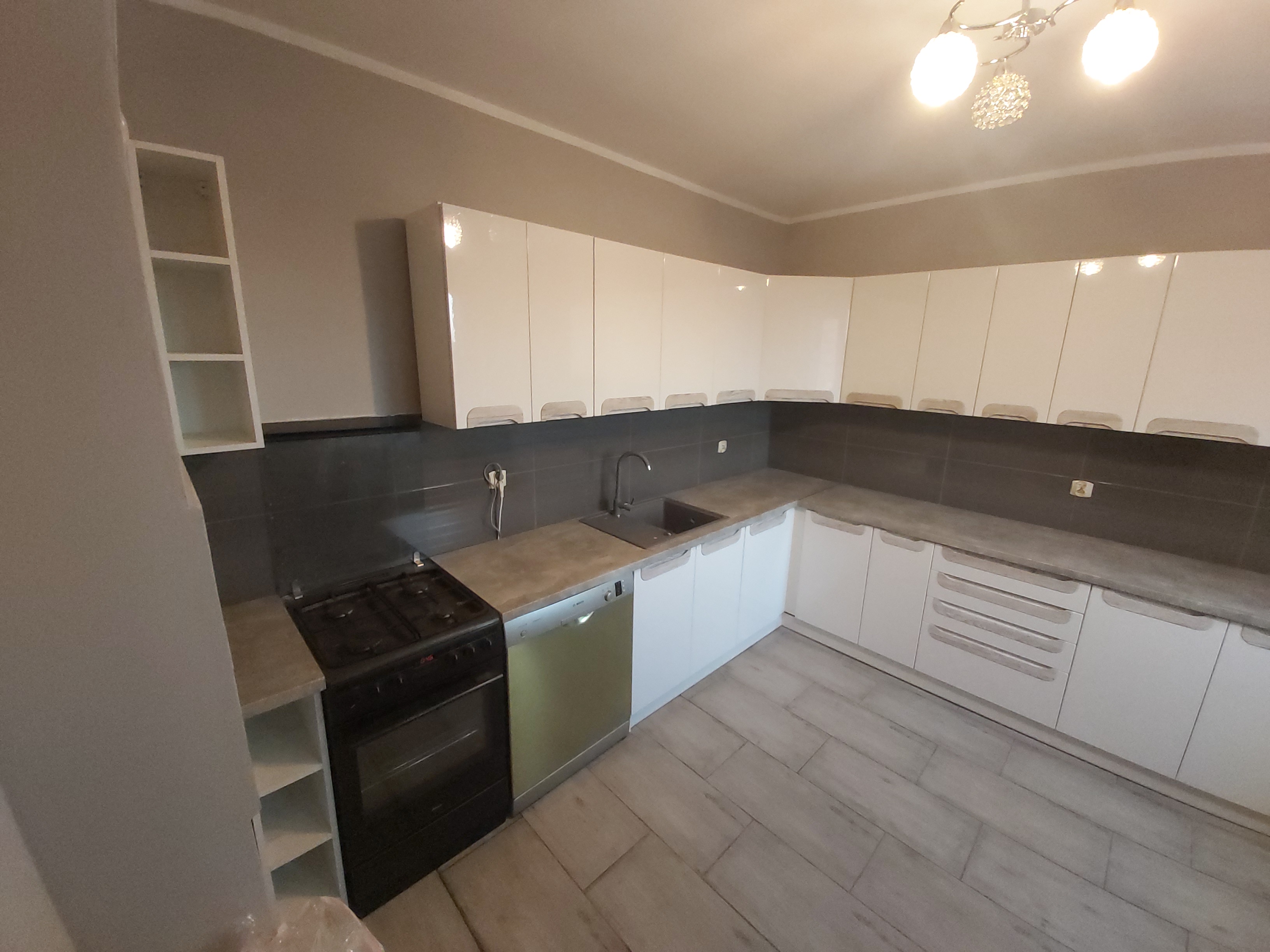 Nowoczesna kuchnia z białymi lakierowanymi szafkami, szarym blatem i ciemnymi płytkami nad blatem, oświetlona lampą sufitową z elementami kryształowymi.