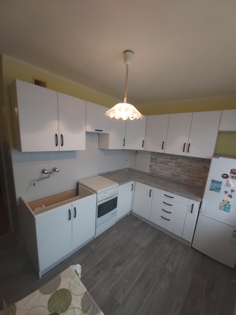 Biała, narożna zabudowa kuchenna z szarym blatem i fragmentem ściany wyłożonej cegiełkami, widoczna na etapie montażu, z niedokończoną szafką pod zlewem i wiszącą lampą z frędzlami.