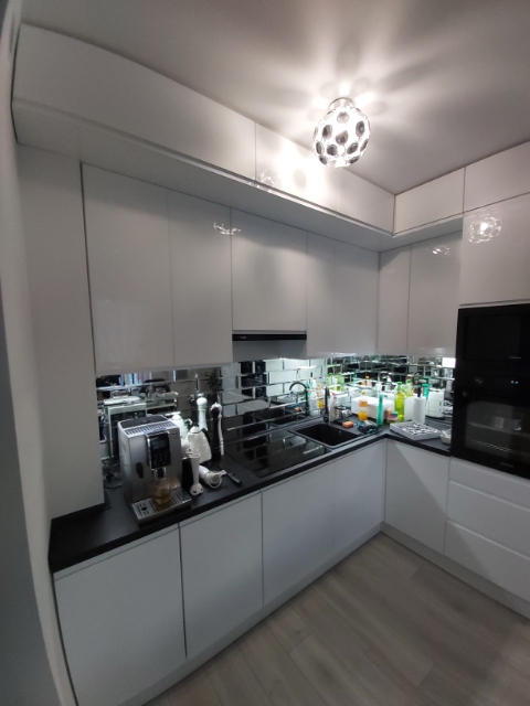 Nowoczesna, biała kuchnia z połyskiem, czarnym blatem i dekoracyjną mozaiką lustrzaną na ścianie. Na blacie ekspres do kawy i akcesoria kuchenne.