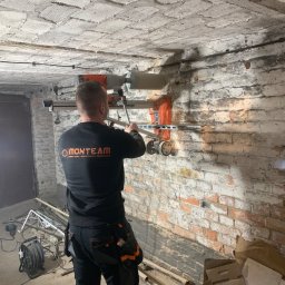 MONTEAM PIOTR POLAK - Pracownik firmy MONTEAM w czarnym ubraniu obsługuje wiertnicę diamentową, montowaną na szynie, podczas wykonywania otworu w starej, ceglanej ścianie piwnicy z nierównym, otynkowanym sufitem.