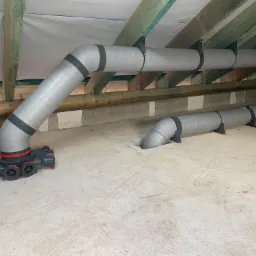 Szare, izolowane rury wentylacyjne z czarnymi uchwytami przymocowane do drewnianej konstrukcji dachu na poddaszu, z widocznym rozdzielaczem z czerwonymi elementami.