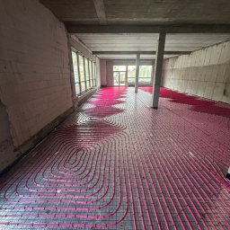 MONTEAM PIOTR POLAK - Różowe rury ogrzewania podłogowego ułożone na szarej macie w surowym wnętrzu z betonowymi ścianami i kolumnami, widok z perspektywy.