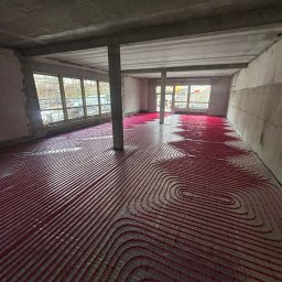 MONTEAM PIOTR POLAK - Czerwone rury ogrzewania podłogowego ułożone w surowym wnętrzu z betonowymi ścianami i kolumnami, widoczne okna z zewnątrz.