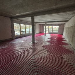 MONTEAM PIOTR POLAK - Czerwone rury ogrzewania podłogowego ułożone na siatce w surowym wnętrzu budynku w trakcie budowy. Widoczne okna i betonowe ściany.