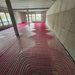 MONTEAM PIOTR POLAK - Czerwone rury ogrzewania podłogowego ułożone na siatce w surowym wnętrzu budynku w trakcie budowy. Widoczne betonowe ściany i słupy.