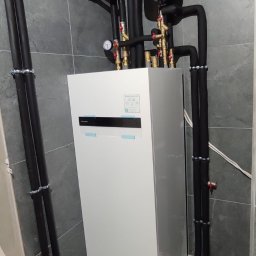 MONTEAM PIOTR POLAK - Biała pompa ciepła Panasonic Aquarea z czarnymi rurami w szarej łazience, widok z przodu. Elementy instalacji hydraulicznej i manometry na górze urządzenia.
