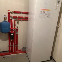 MONTEAM PIOTR POLAK - Biała pompa ciepła Atlantic Alfea z niebieskim zbiornikiem wyrównawczym i czerwonymi rurami instalacji grzewczej w surowym pomieszczeniu z betonową posadzką.