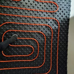 MONTEAM PIOTR POLAK - Pomarańczowe rury ogrzewania podłogowego ułożone na czarnej macie izolacyjnej z wypustkami; widoczny fragment buta montera.