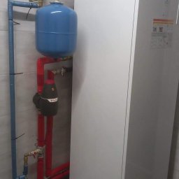 MONTEAM PIOTR POLAK - Nowoczesna, biała pompa ciepła z niebieskim zbiornikiem wyrównawczym i czerwonymi rurami w pomieszczeniu z szarą glazurą.