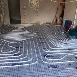 MONTEAM PIOTR POLAK - Białe rury ogrzewania podłogowego ułożone na czarnej siatce, z widocznymi narzędziami i rozdzielaczem w tle, w surowym wnętrzu z wystającymi kablami.