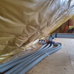 Szare, elastyczne rury wentylacyjne podłączone do rozdzielacza w nowym, ocieplonym poddaszu, widoczne mocowania i folia izolacyjna.