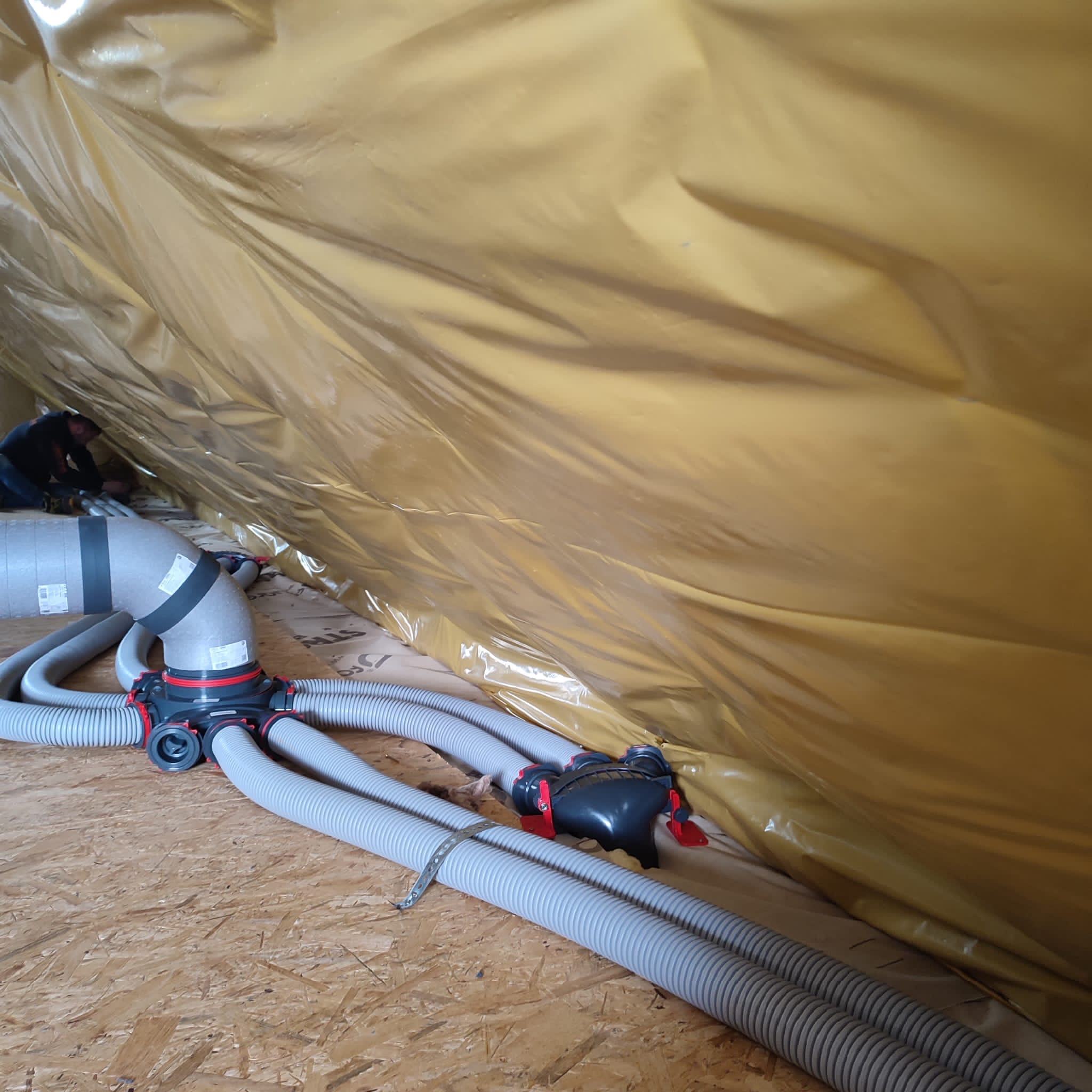 Szare, karbowane przewody wentylacyjne podłączone do rozdzielacza w pomieszczeniu z izolacją ze złotej folii, widoczny fragment pracującej osoby w tle.