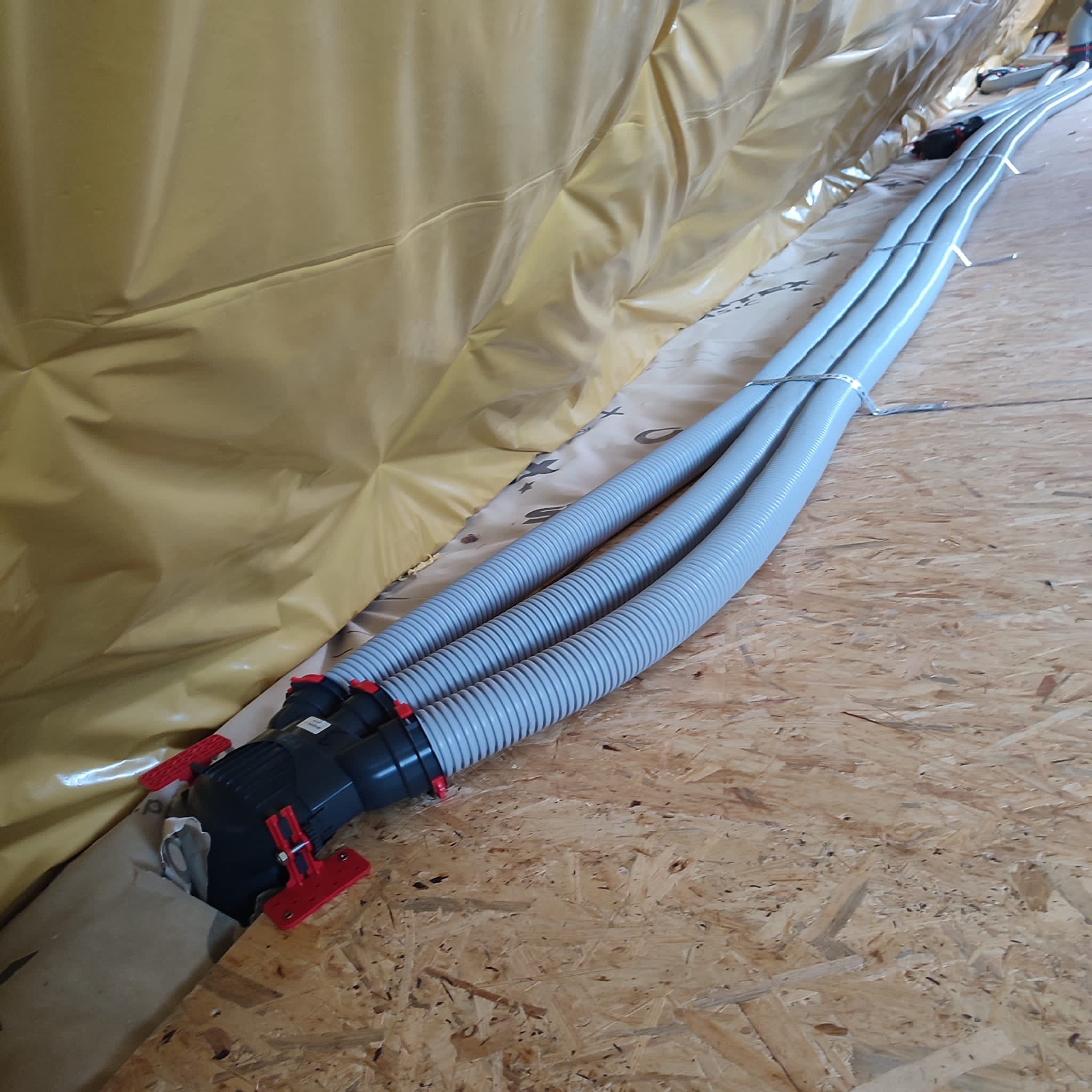 Szare, karbowane rury wentylacyjne połączone czarnym rozdzielaczem z czerwonymi zaciskami, ułożone na płycie OSB, z folią izolacyjną w tle.