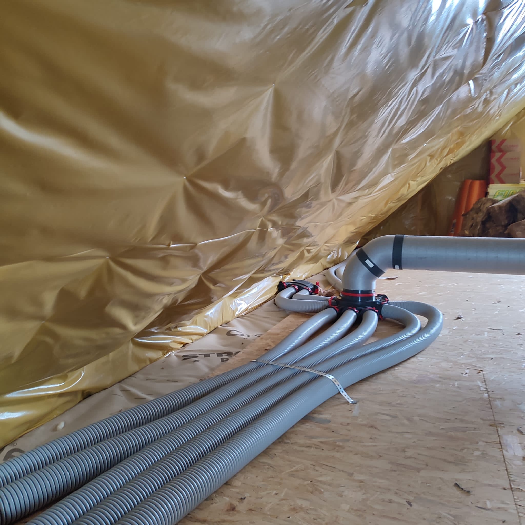 Szare, elastyczne rury wentylacyjne podłączone do rozdzielacza w nowym, ocieplonym poddaszu, widoczne mocowania i folia izolacyjna.