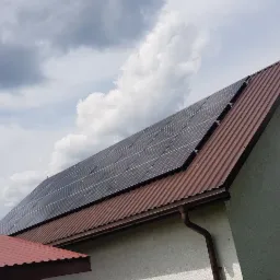 Panel fotowoltaiczny na brązowym, blaszanym dachu domu jednorodzinnego, widok z dołu na tle pochmurnego nieba.